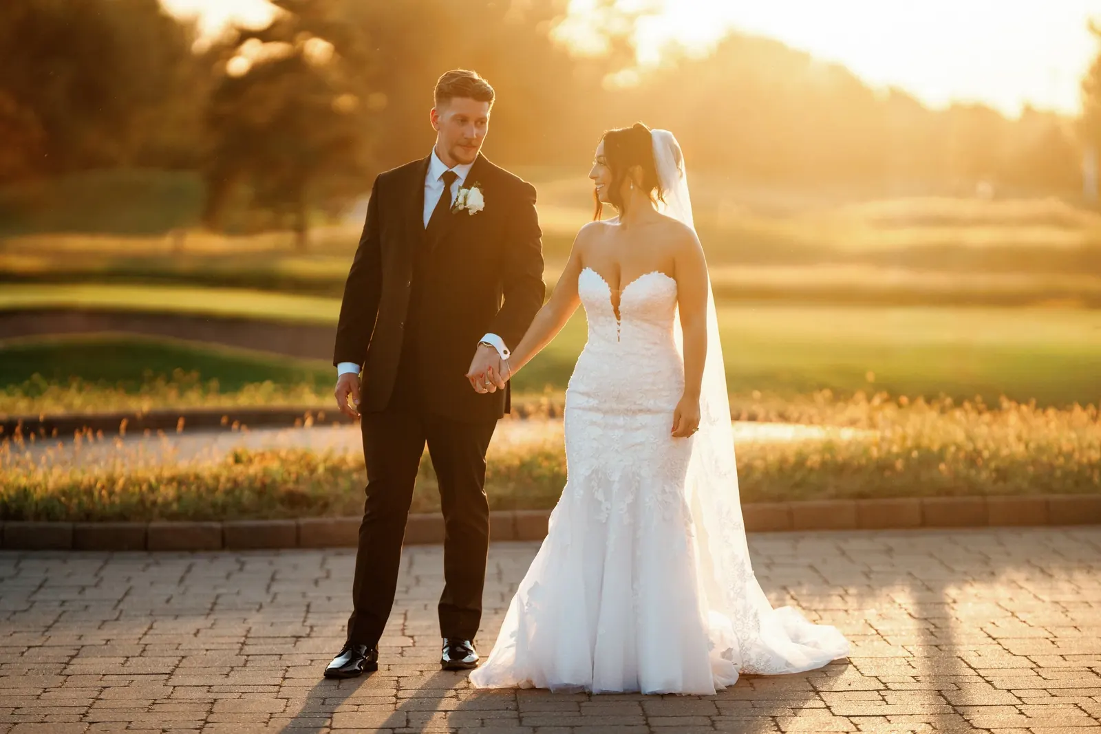 A bride and groom holding hands on a stone path, lit by the setting sun